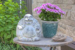 Das Bild zeigt einen kleinen lächlenden Buddha aus hellgrauem Steinzeug. Er steht auf einem kleinen runden Tisch mit einer grauen Steinplatte. Neben dem Tisch befindet sich eine kleine Pflanze mit rosafarbenen kleinen Blüten und grünen kleinen Blättern. Vor der Pflanze liegt die Skulptur einer Rosenblüte aus Steinzeug. Hinter dem Tisch sieht man rechts eine grüne Buschhortensie und links eine hellbeige Steinmauer aus Naturstein.