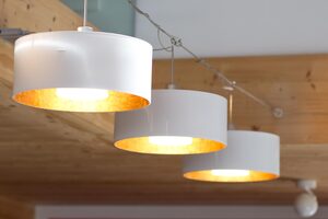 Das Beitragsild zu meinem Blogbeitrag „Raumbeleuchtung nach klassischem Feng Shui". Auf dem Bild siehst du drei runde weiße Leuchten, die an einem Seil hängen, dass in einer Galerie gespannt ist. Das Seil ist in Holzbalken verankert. Die drei Leuchten bestehen aus einem äußeren gläsernen Schirm mit Durchmesser ca. 30 Zentimeter, innen vergoldet. In der Mitte hängt ein kleiner weißer Schirm, mit Durchmesser ca. 12 Zentimeter. Die drei Leuchten strahlen ein sehr warmes Licht nach unten ab.