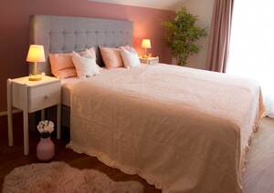 Ein modernes Schlafzimmer mit rosa-grauer Farbgebung. In der Mitte befindet sich ein Boxspringbett mit rosafarbener Tagesdecke, im Hintergrund ist eine grüne Zimmerpflanze. Links und rechts neben dem Bett stehen Nachttische mit kleinen Leuchten, die ein gelbwarmes Licht abstrahlen. Auf dem Bett sind rosafarbene und weiße Kissen. Vor dem Bett liegt ein rosafarbener kleiner Plüschvorleger. Vor dem Nachttisch auf der linken Seite steht eine rosafarbene Vase mit rosafarbenen Tulpen.