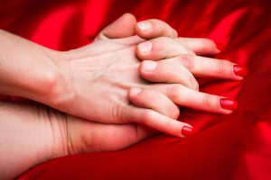 Das Titelbild des Beitrags "Sexualität und Feng Shui: Ein aufregender Weg zur harmonischen Erfüllung". Das Bild zeigt zwei verschlungene Hände auf einer intensivroten Decke.