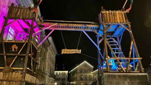 Ein Bild vom nächtlichen Mittelaltermarkt in Esslingen am Neckar, auf dem das Eingangstor aus zwei hölzernen Türmen sichtbar ist. Die Türme sind pink und blau angestrahlt.