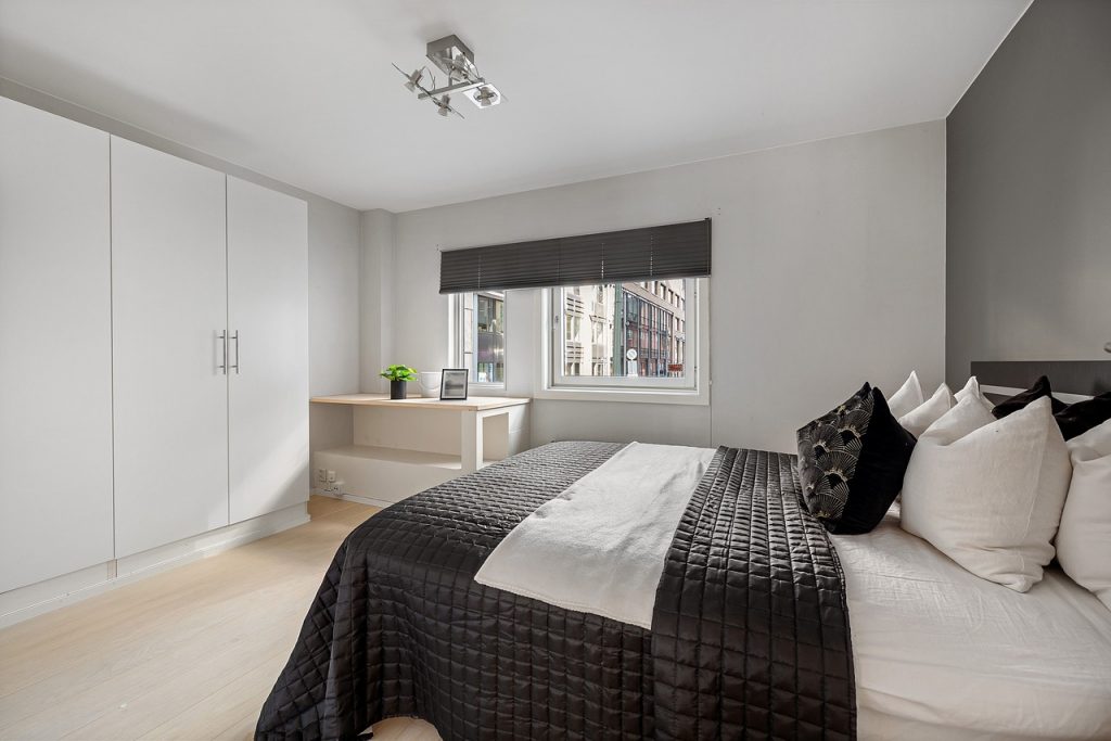 Ein modernes Schlafzimmer in den Farben Weiß und einem modernen dunklen Grauton gestaltet. Auf der rechten Seite steht ein Bett mit einer schwarzen Decke mit Quadraten. Auf dem Bett befinden sich vier weiße und zwei schwarze Kissen. An der linken Seite des Raumes steht ein geschlossener Kleiderschrank mit länglichen dünnen Metallgriffen (ca. 20 cm lang). Im Hintergrund befindet sich ein Fenster mit Blick auf eine Häuserfront in einer Innenstadt. Vor dem Fenster befindet sich ein Tisch mit einer Pflanze, einem Blumentopf und einem Bild.