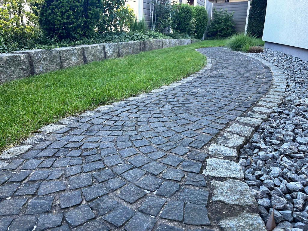 Ein modern gestalteter Weg aus Basaltpflaster, eingefasst in graue Einzeiler aus Granit. Der Weg ist ganz bewusst mit Rundungen entworfen, um den Energiefluss zu optimieren.