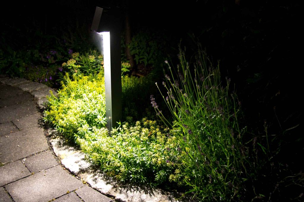 In der Bildmitte eine graue Gartenleuchte bei Nacht. Die Leucht ist etwa 1 m hoch und strahlt nach unten, sodass man nicht geblendet wird. Auf dem Bild sieht man Pflanzen im Licht und eine geschwungene Hofeinfassung aus Granitpflaster. Der Hofbelag besteht aus grauem Betonpflaster.