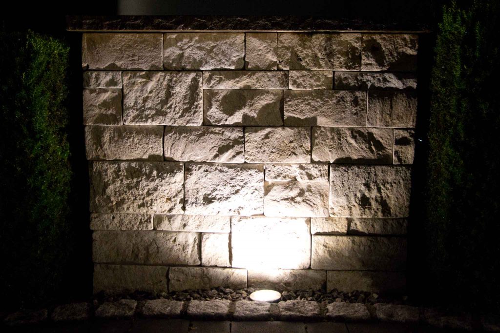 Auf dem Bild siehst du eine Natursteinwand aus Kalksteinen gemauert, welche durch eine Bodenleuchte angestrahlt wird. Links und rechts der Mauer kannst Du noch grüne Hecken erkennen. Die Mauer ist an der Unterseite durch eine Pflasterzeile aus Granit abgegrenzt, der Zwischenraum ist mit Kalksplitt gefüllt.