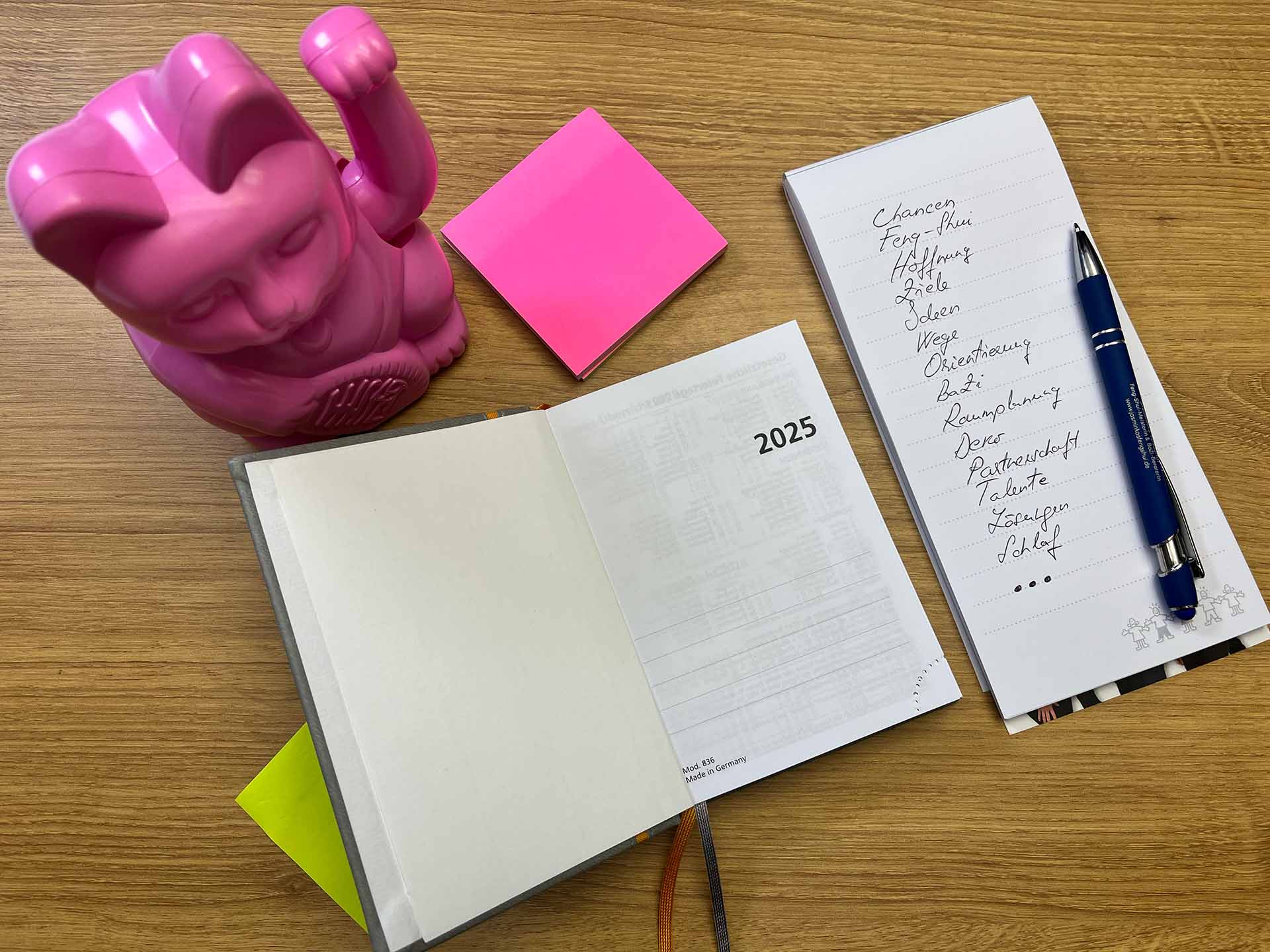 Auf dem Bild siehst Du in der Mitte ein geöffnetes Buch mit einem Jahreskalender für 2025. Rechts daneben befindet sich ein Schreibblock mit einem blauen Stift. Auf dem Block befinden sich folgende Stichworte geschrieben: Chancen, Feng Shui, Hoffnung, Ziele, Ideen, Wege, Orientierung, BaZi, Raumplanung, Deko, Partnerschaft, Talente, Lösungen, Schlaf und drei Punkte. Über dem Kalenderbuch befindet sich ein quadratischer Post-it-Block in pinker Farbe. Links daneben siehst du eine pinkfarbene chinesische Winkekatze aus Kunststoff. Die Gegenstände befinden sich auf einer Schreibtischplatte aus Eichenholz.