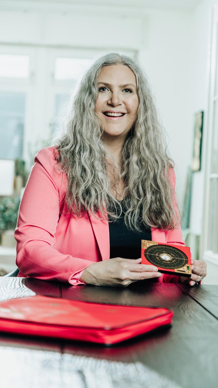 Jasminka Hepperle, an einem schwarzen Schreibtisch sitzend. Sie trägt einen rosafarbenen Blazer und darunter einen schwarzen Pullover. In der Hand hält sie einen kleinen Luo Pan Kompass, davor liegt eine rote Hülle, in welcher sich ein großer Luo Pan Kompass befindet.