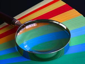 Eine Lupe liegt auf leuchtend regenbogenfarbenem Papier in Nahaufnahme.
