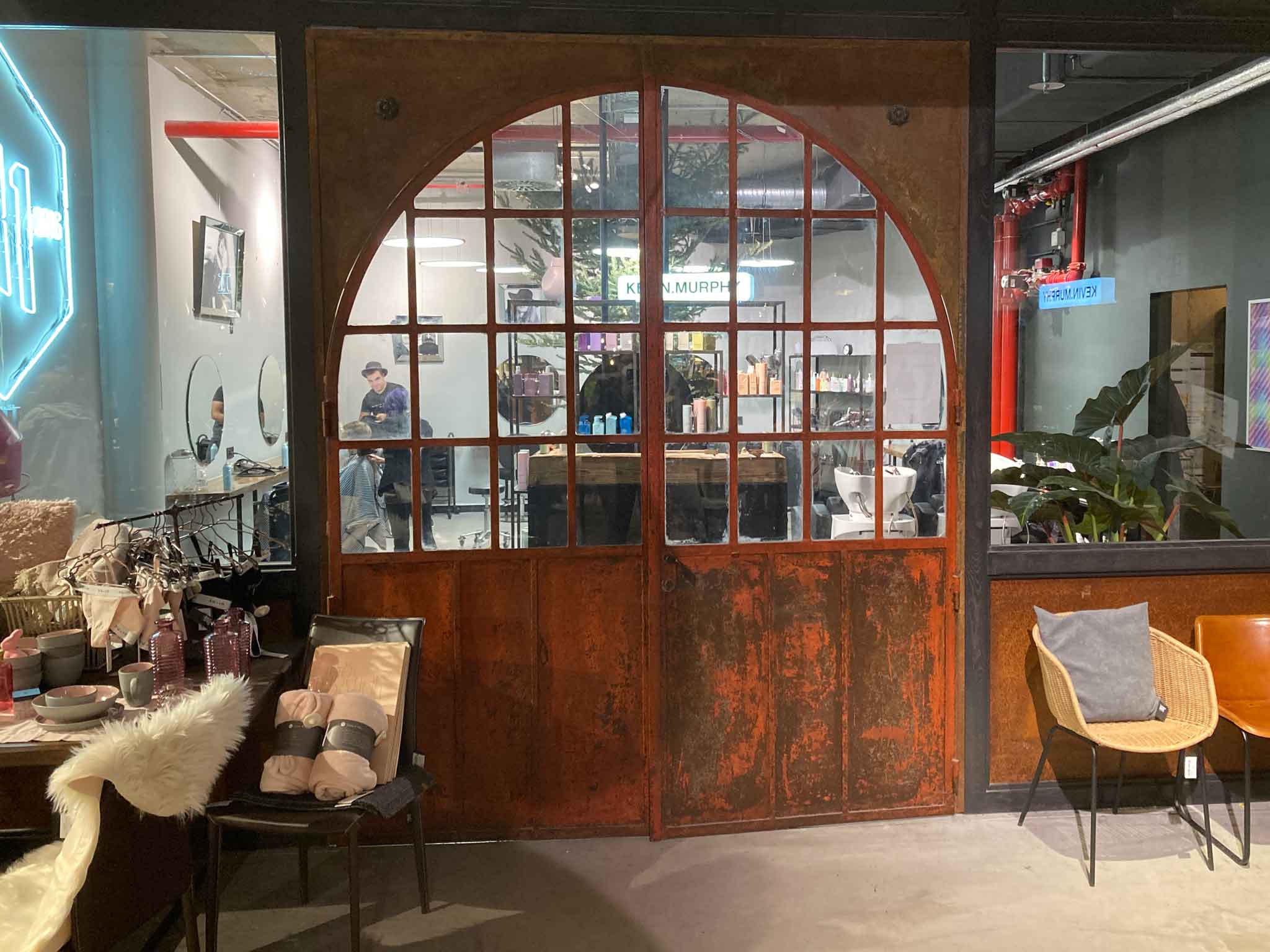 Blick auf eine alte, bogenförmige Eingangstüre vor einem modernen Friseursalon. Die Tür ist zweiflüglig, die oberen zwei Drittel sind gitterförmig verglast. Die Tür ist orange eingefärbt in altem rostfarbenem Stil. Im Vordergrund sieht man rechts zwei Stühle und links Dekogegenstände, die zum Verkauf stehen.