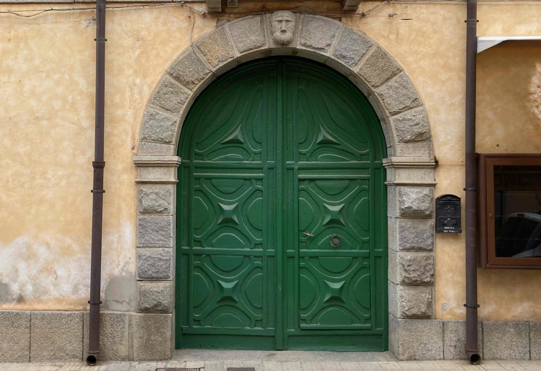 Beispiel für eine historische Eingangstüre. Die grüne massive Holztüre ist zweiflüglig und hat oben einen Rundbogen. Die Tüe ist mit grau-braunen grob behauenen Natursteineine eingefasst. Ganz oben befindet sich ein Gesicht, das aus dem Naturstein herausgearbeitet wurde.
