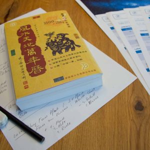 Die Ausarbeitung einer BaZi-Analyse erfordert fundiertes Fachwissen. Auf dem Bild siehst Du notwendige Unterlagen wie handschriftliche Notizen, ein Buch mit dem chinesischen Kalender (Jahre 1864 bis 2100) und ein Beispiel für eine fertige Analyse.