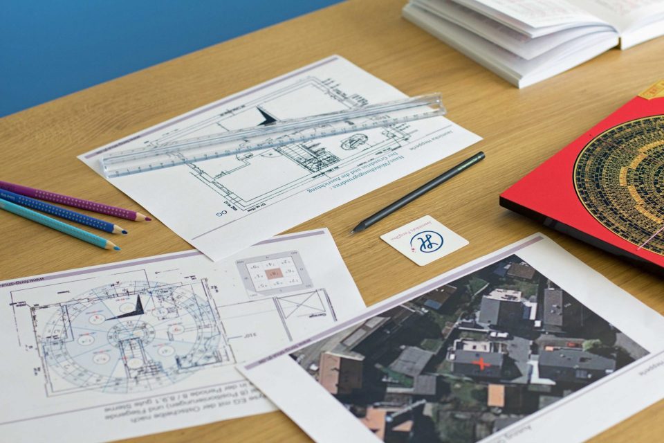 Du siehst einen eichenfarbigen Schreibtisch, auf dem sich Gegenstände befinden, welche bei einer Feng Shui Beratung zum Einsatz kommen: Pläne, Lineal, Bleistift, Luo Pan Kompass. Im Hintergrund sieht man eine blaue Wand.