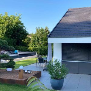 Ein modernes Wohnhaus mit einem streng formal gestalteten Garten. Im Vordergrund sieht man eine Terrasse mit grauen großformatigen Platten und im Hintergrund links einen Pool. Als Dekoelemente sind moderne Leuchen und Kugeln aus Edelstahl eingesetzt.